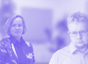 Two colleagues stand in a bright office—woman with glasses on the left, man with glasses on the right, blurred background behind them.