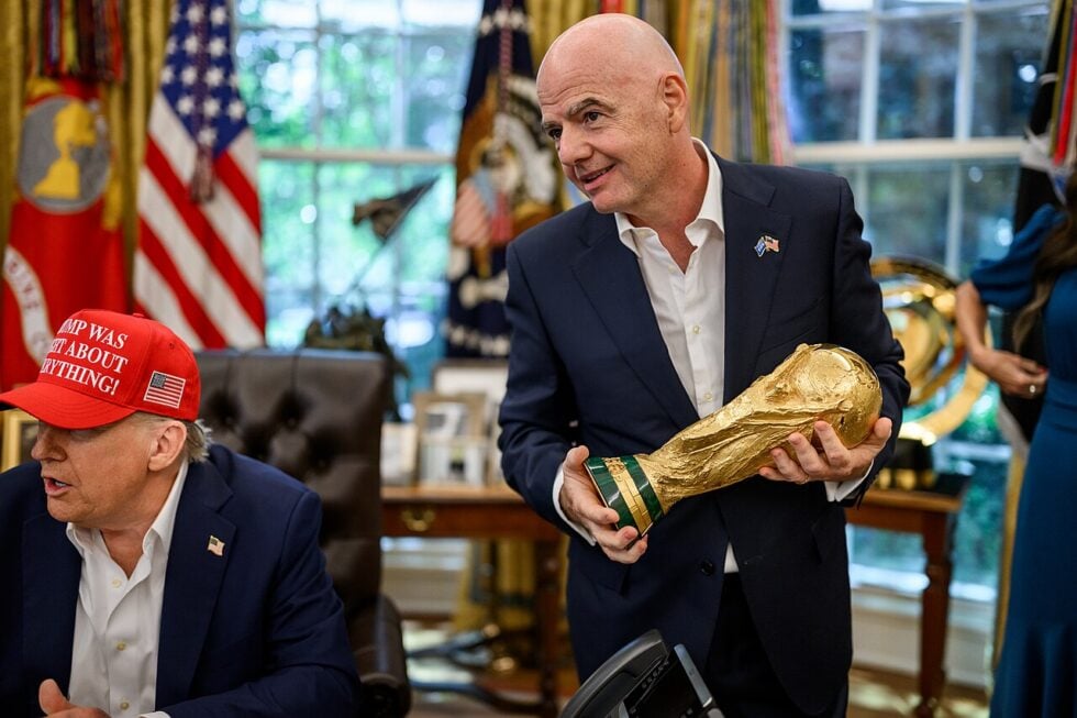 Man in a dark suit holds a large gold trophy in an office with American flags in the background.