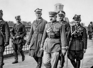 Group of historical military officers in dress uniforms stride along a railing-lined promenade, medals and sashes visible on their chests