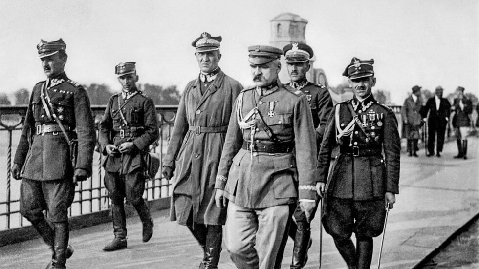 Group of historical military officers in dress uniforms stride along a railing-lined promenade, medals and sashes visible on their chests