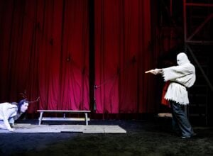 Theatrical scene onstage with red curtains; a masked performer in white points an orange-tipped prop gun at a kneeling figure on the ground.