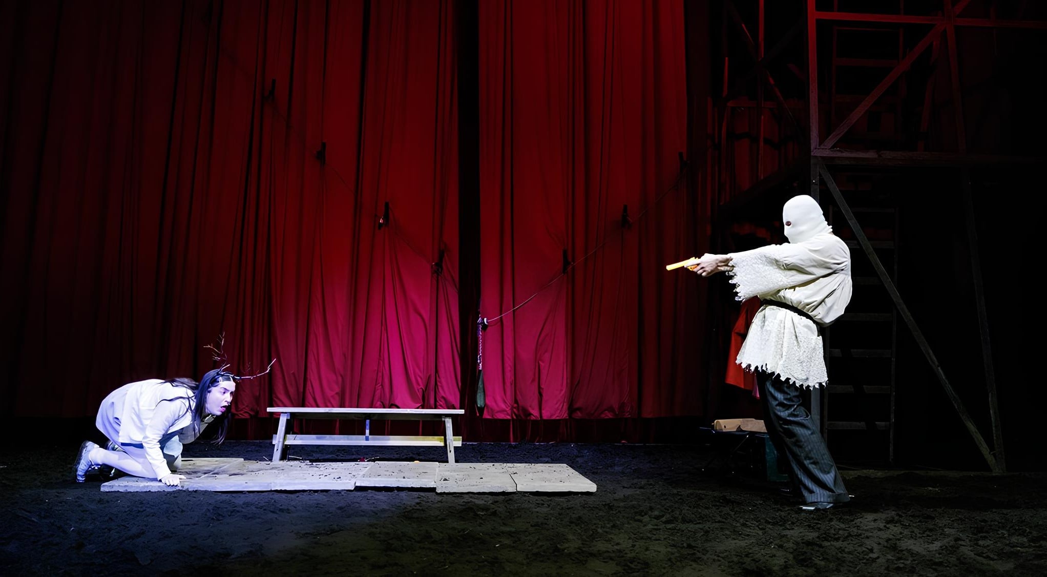 Theatrical scene onstage with red curtains; a masked performer in white points an orange-tipped prop gun at a kneeling figure on the ground.