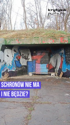 Podziemny przejazd/klatka z graffiti na ścianach i zamkniętą bramą, otoczoną sypiącymi się cegłami i leżącymi deszkami.