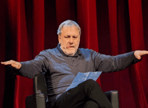 Speaker on a stage with red curtains, arms outstretched and a paper in hand while presenting.