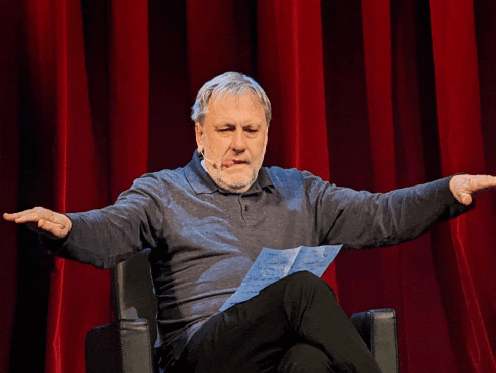 Speaker on a stage with red curtains, arms outstretched and a paper in hand while presenting.