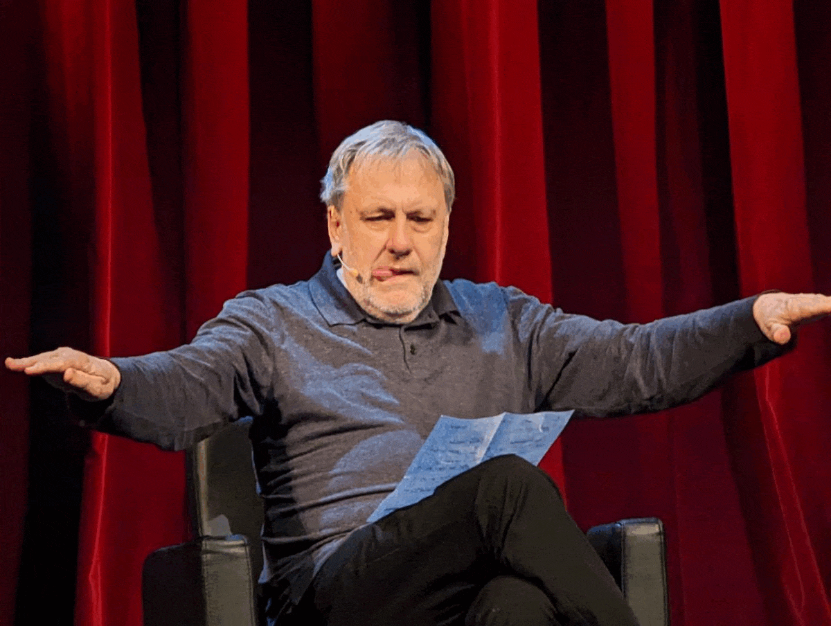 Speaker on a stage with red curtains, arms outstretched and a paper in hand while presenting.