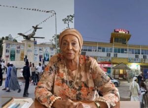 Elderly woman in a floral outfit and headwrap sits at a desk in a busy town square, with an airplane statue and hotel in the background.