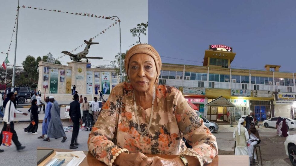Elderly woman in a floral outfit and headwrap sits at a desk in a busy town square, with an airplane statue and hotel in the background.