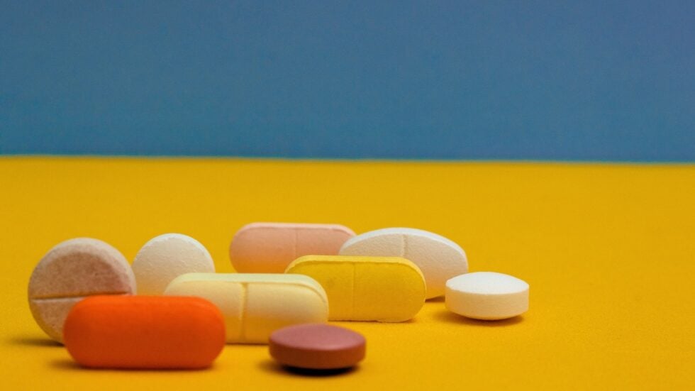 Assorted colorful pills and tablets scattered on a yellow surface with a blue wall in the background.