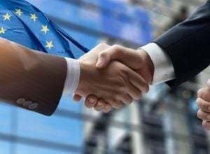 Two people in business suits shake hands with an EU flag in the background, signaling a European agreement or partnership.