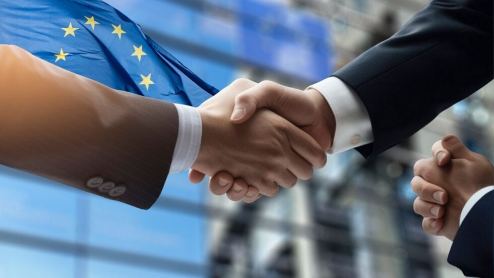 Two people in business suits shake hands with an EU flag in the background, signaling a European agreement or partnership.