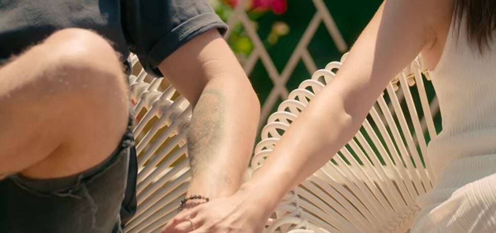 Close-up of two people holding hands while sitting on a white woven outdoor chair, sunlight highlighting a tattooed forearm on the left.