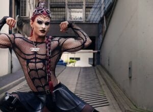 Drag performer striking a strong bodybuilding pose in a black mesh bodysuit and leather skirt in an urban alleyway