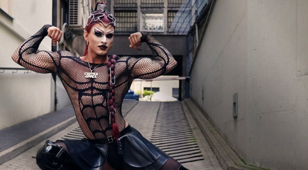Drag performer striking a strong bodybuilding pose in a black mesh bodysuit and leather skirt in an urban alleyway