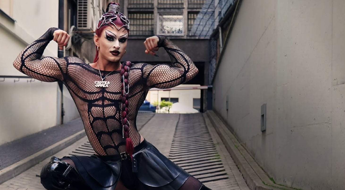 Drag performer striking a strong bodybuilding pose in a black mesh bodysuit and leather skirt in an urban alleyway