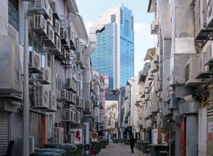 Narrow alley lined with many wall-mounted air conditioners; a person walks toward a blue glass skyscraper in the distance, with bins along the sides.