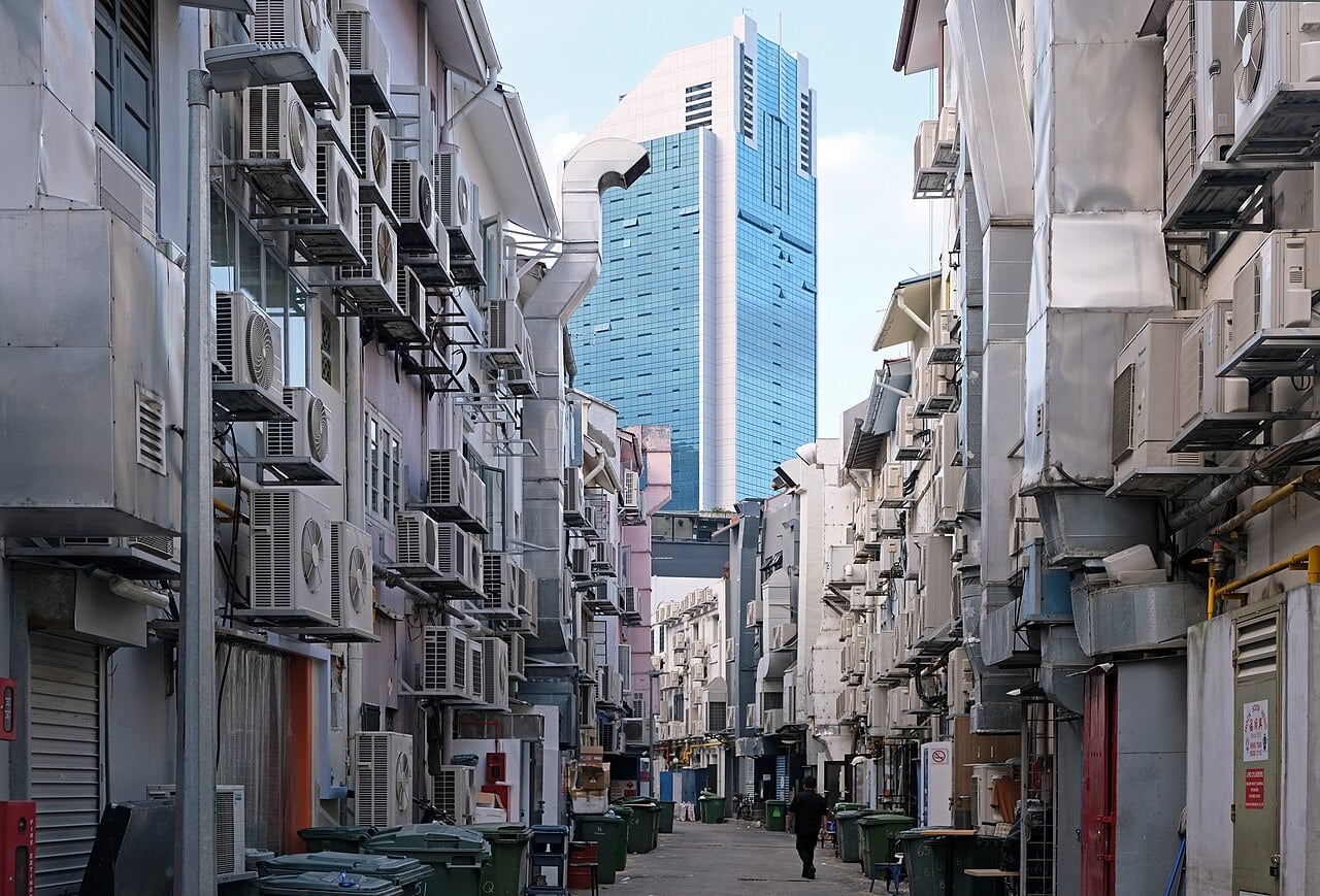Narrow alley lined with many wall-mounted air conditioners; a person walks toward a blue glass skyscraper in the distance, with bins along the sides.