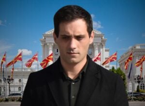 Man in a dark coat stands in front of a white neoclassical building with red-yellow flags and a blue sky in the background.