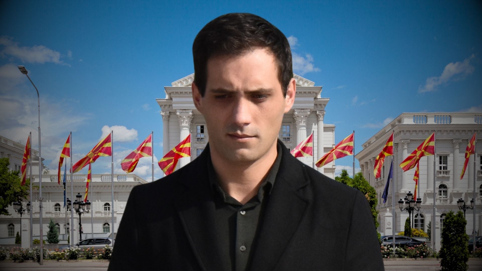 Man in a dark coat stands in front of a white neoclassical building with red-yellow flags and a blue sky in the background.