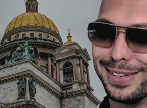 Partial image showing a man wearing dark sunglasses smiling on the right, beside a historic building with a large gold dome and sculpted statues on the left.
