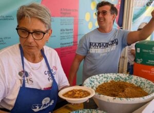 Two volunteers at a food stall presenting bowls of cooked food, with colorful banners in the background.