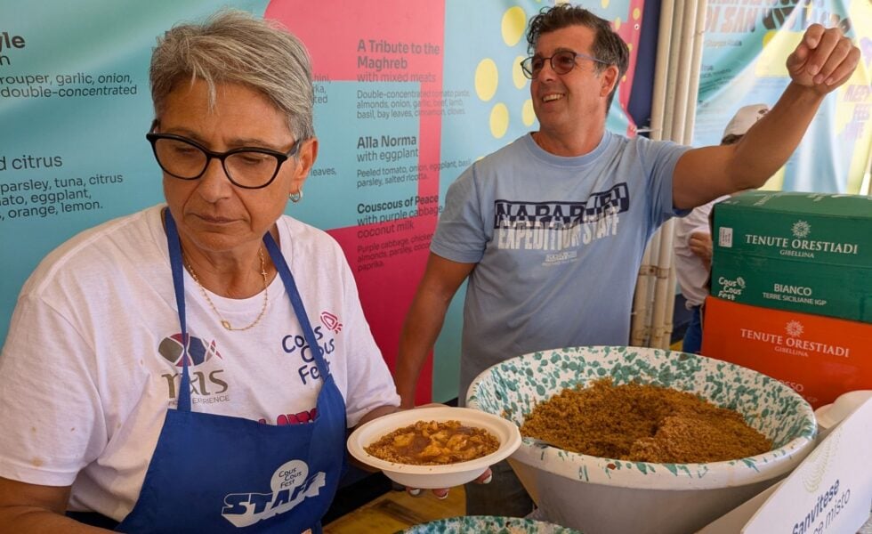 Two volunteers at a food stall presenting bowls of cooked food, with colorful banners in the background.