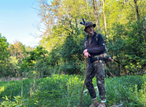 Hiker with a trekking pole stands in a sunlit forest clearing wearing a hat, backpack, and camouflage pants.