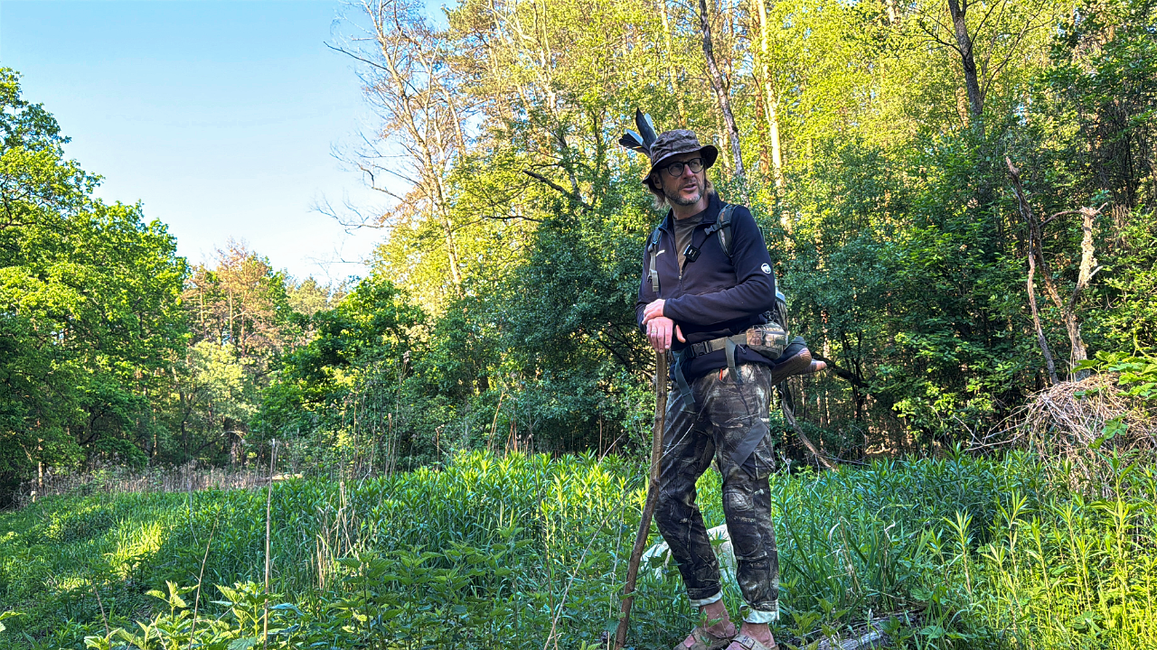 Hiker with a trekking pole stands in a sunlit forest clearing wearing a hat, backpack, and camouflage pants.