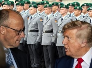 Two men in suits conversing in the foreground with a line of uniformed soldiers in gray uniforms and green berets behind them