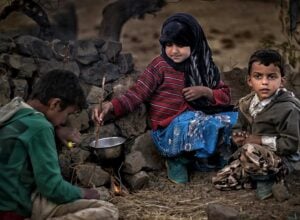 Three children sit around a small outdoor fire, cooking in a metal pot beside a stone wall.