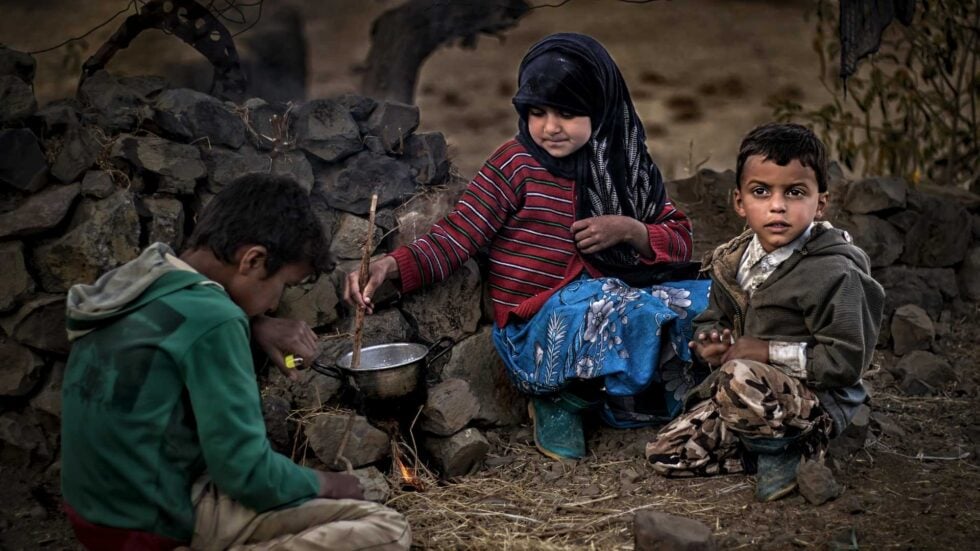Three children sit around a small outdoor fire, cooking in a metal pot beside a stone wall.