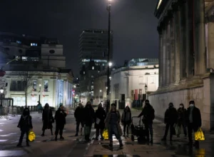 Group of people wearing masks standing on a city square at night, holding glowing yellow lanterns.