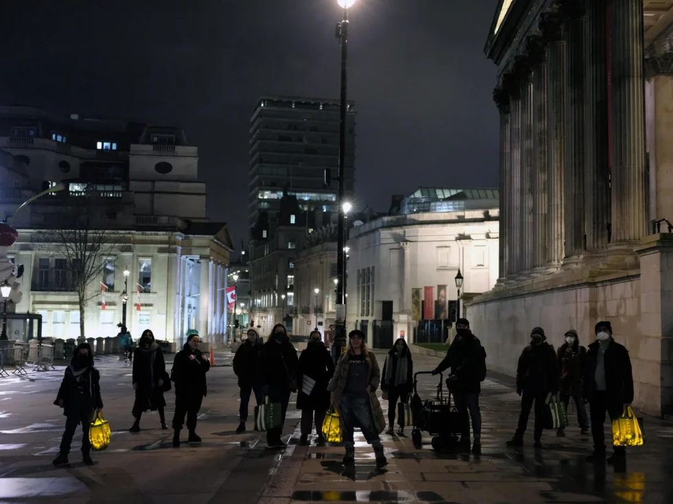 Group of people wearing masks standing on a city square at night, holding glowing yellow lanterns.