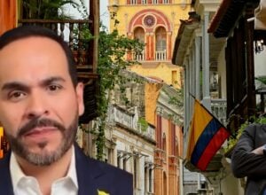 Two men in suits in a colorful colonial street; left foreground close-up, right man smiling with a Colombian flag visible in the scene.