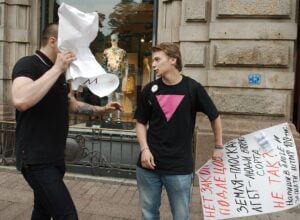 Two men on a city sidewalk; one hides his face with a crumpled sheet of paper while the other talks, with a large handwritten sign nearby.
