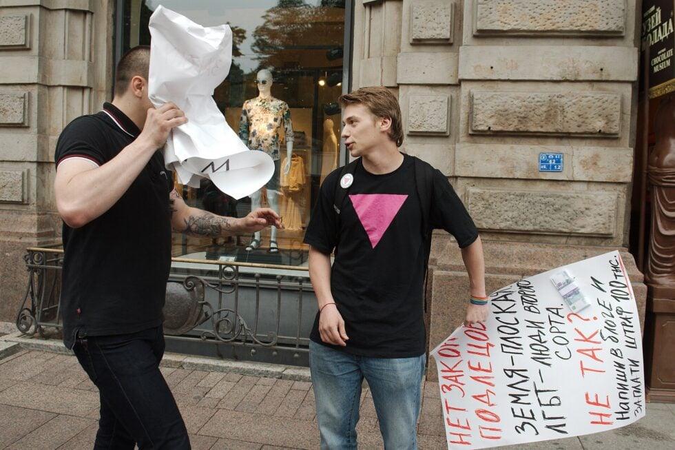 Two men on a city sidewalk; one hides his face with a crumpled sheet of paper while the other talks, with a large handwritten sign nearby.