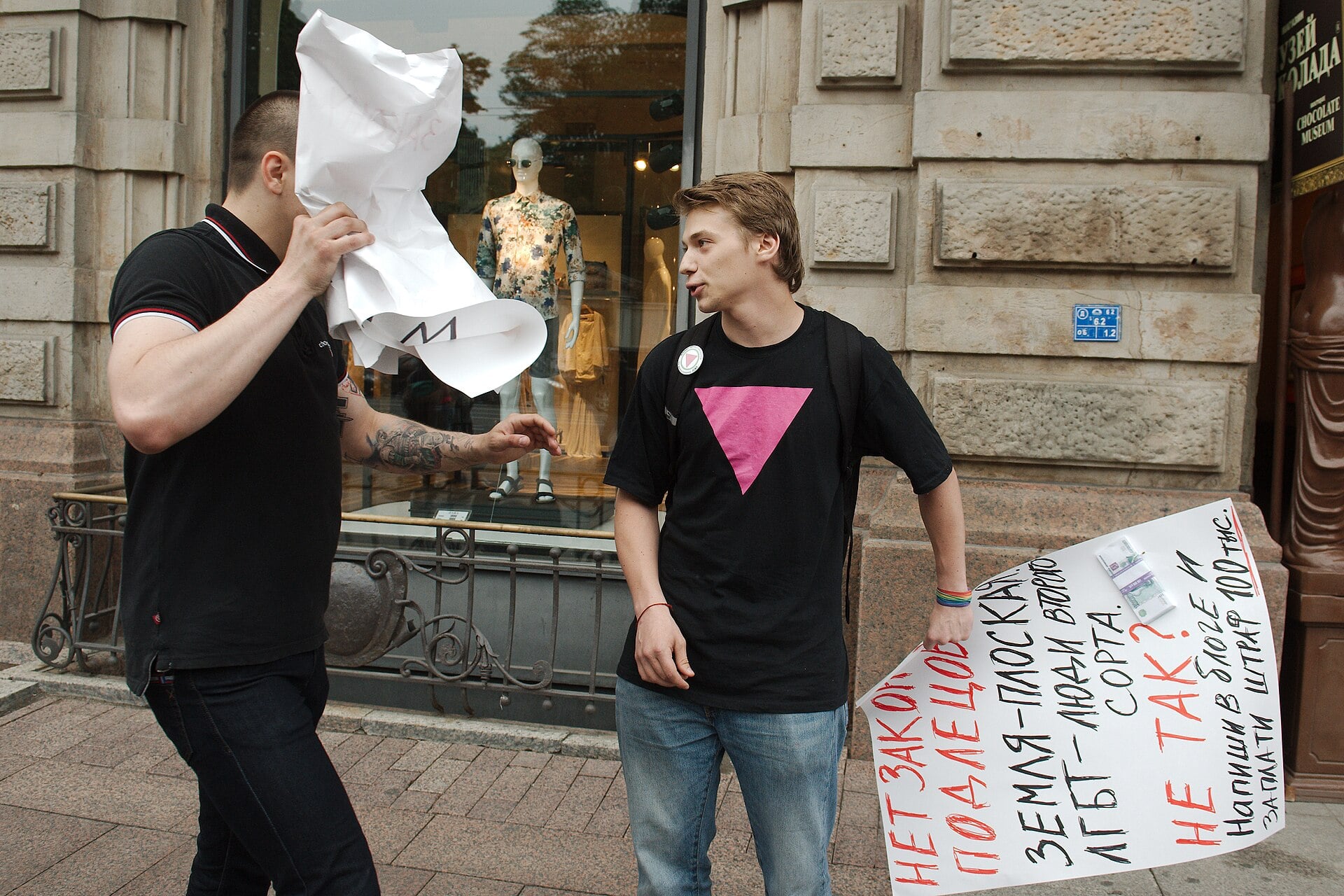 Two men on a city sidewalk; one hides his face with a crumpled sheet of paper while the other talks, with a large handwritten sign nearby.