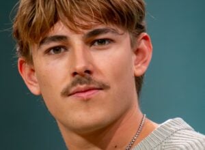 Close-up of a young man with light brown hair and a silver chain, wearing a striped knit sweater against a teal background.