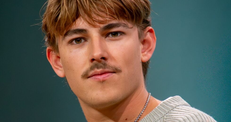 Close-up of a young man with light brown hair and a silver chain, wearing a striped knit sweater against a teal background.