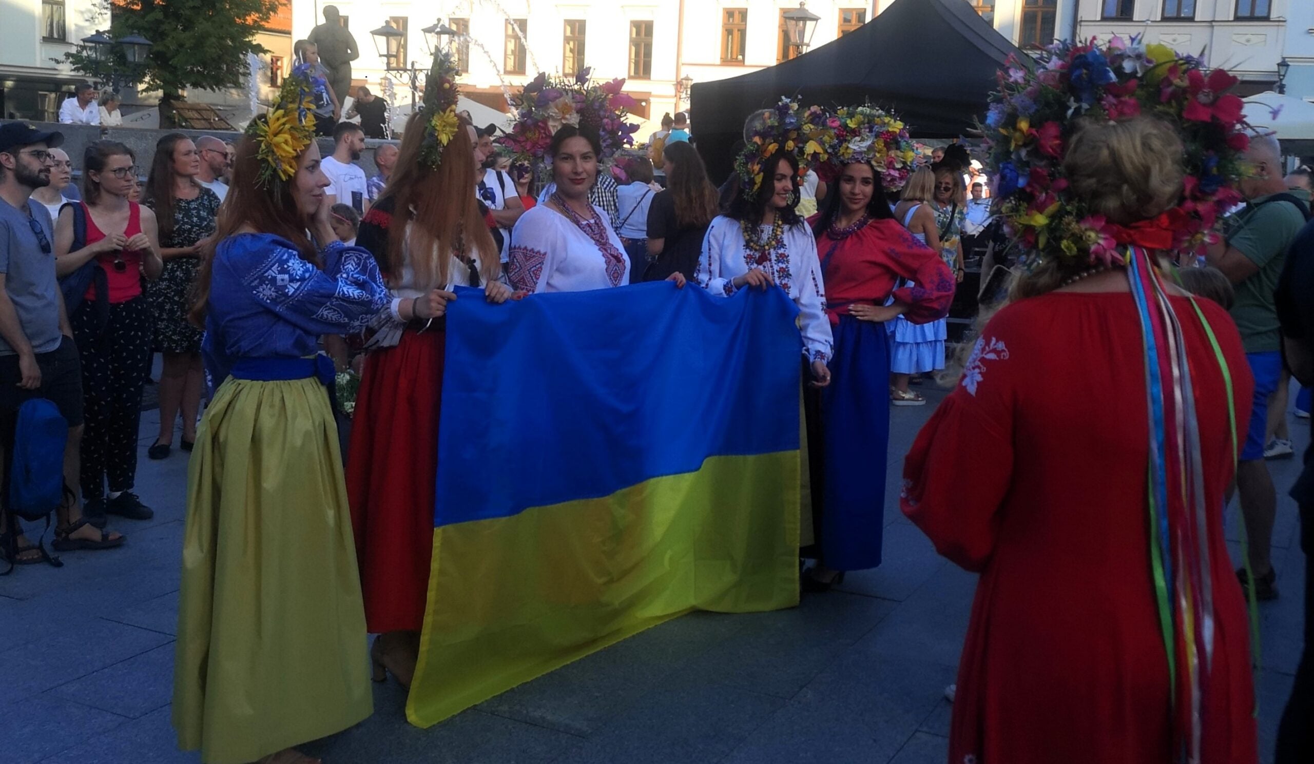 Group of people in traditional Ukrainian dress holding a large blue-and-yellow flag at a festive street event.