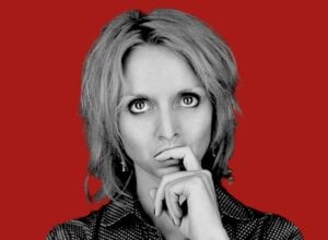 Portrait of a woman in black-and-white, with a contemplative pose and hands near her mouth against a red background.