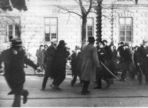 Black-and-white street parade with blurred crowds in coats and hats; a stone building in the background.