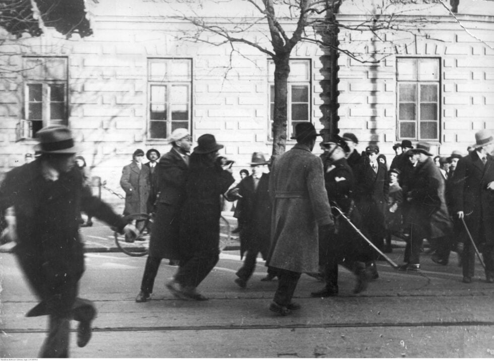 Black-and-white street parade with blurred crowds in coats and hats; a stone building in the background.