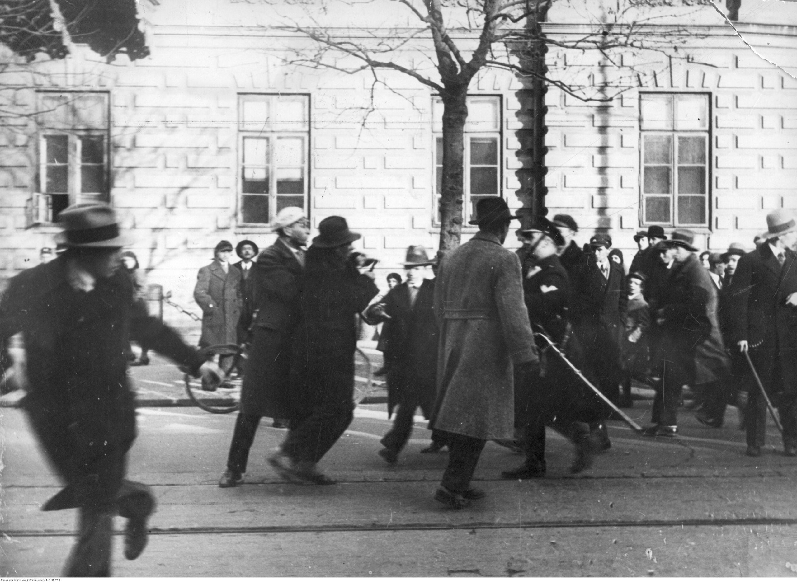 Black-and-white street parade with blurred crowds in coats and hats; a stone building in the background.