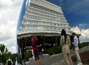 Curved, multi-level white hotel or condo building with glass balconies, set against a blue sky; people stand and walk in a plaza in the foreground.