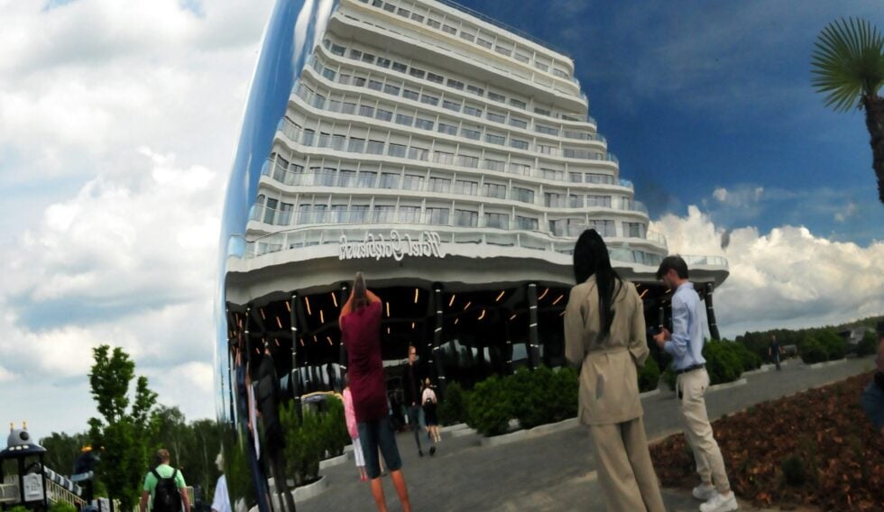 Curved, multi-level white hotel or condo building with glass balconies, set against a blue sky; people stand and walk in a plaza in the foreground.