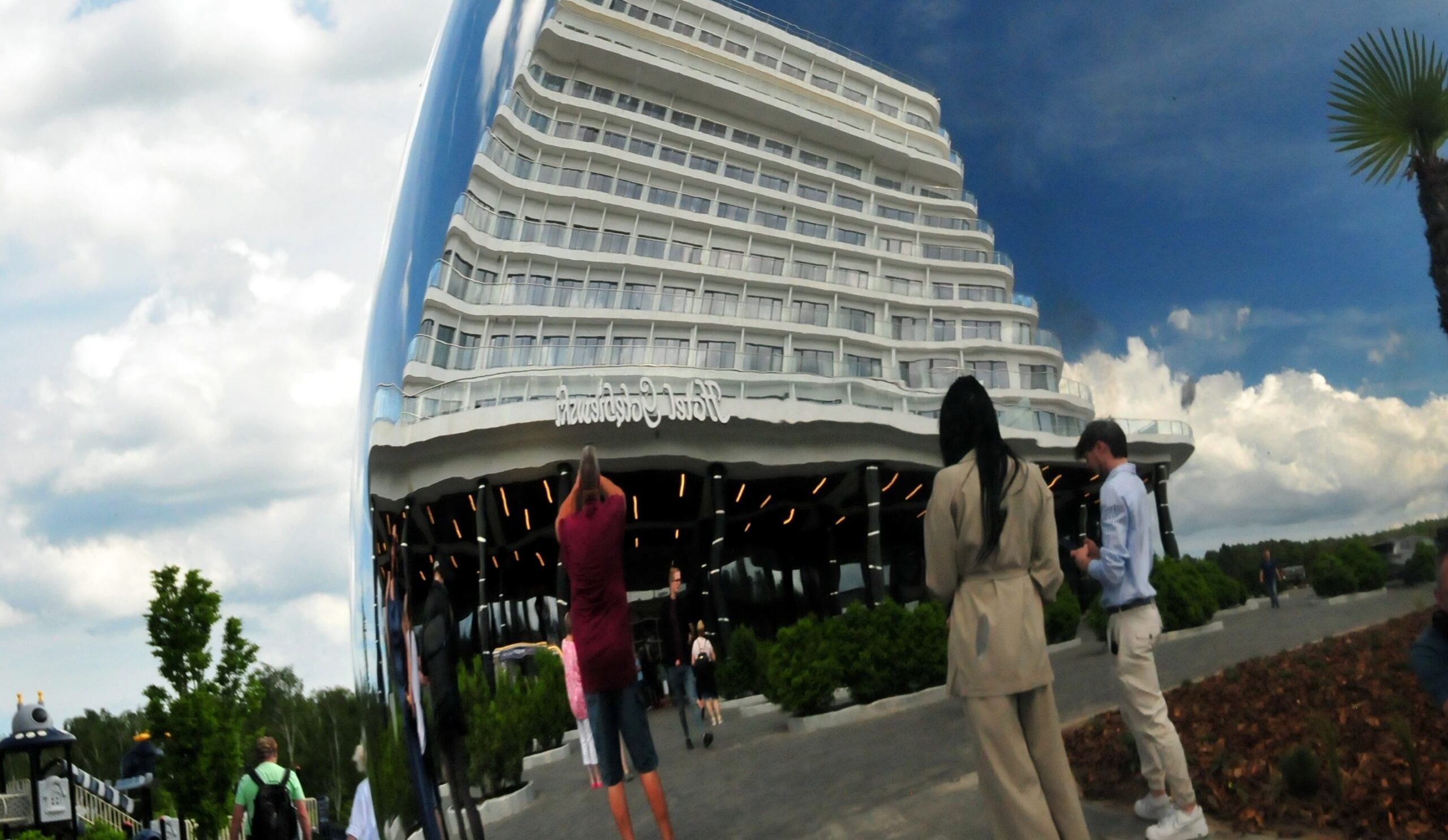 Curved, multi-level white hotel or condo building with glass balconies, set against a blue sky; people stand and walk in a plaza in the foreground.
