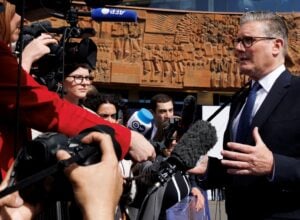 Politician in a dark suit speaks to a cluster of reporters holding microphones and cameras outdoors in front of a carved orange wall.