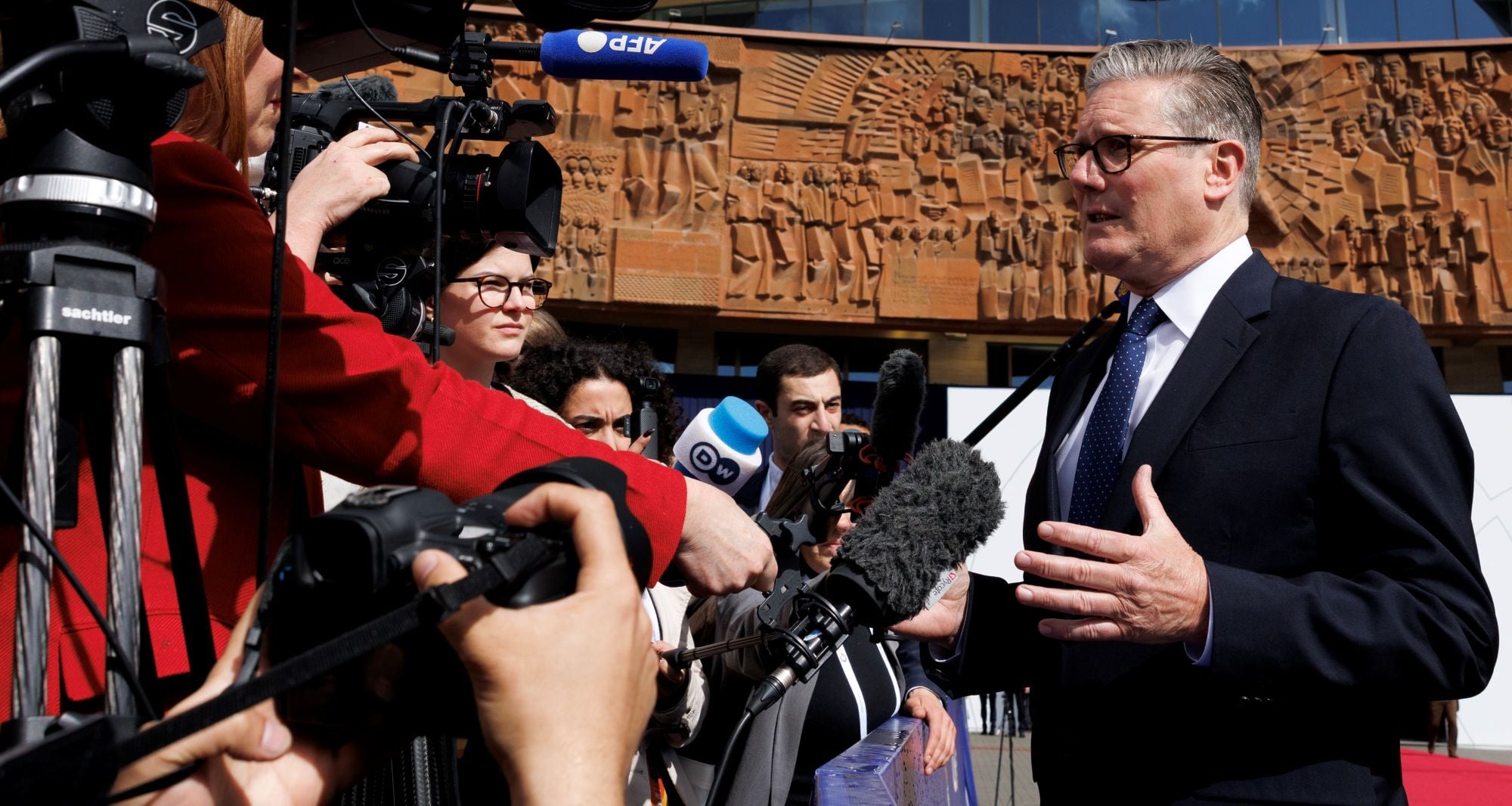 Politician in a dark suit speaks to a cluster of reporters holding microphones and cameras outdoors in front of a carved orange wall.