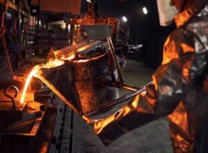 Molten metal pouring into a mold in a steel foundry, illuminated by orange glow and industrial equipment nearby.